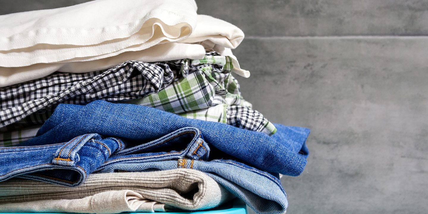 Folded bedding, jeans, towels on a blue box against the blurred background of a gray concrete wall in the bathroom. Pile of laundry and clothes prepared for washing. Closeup view