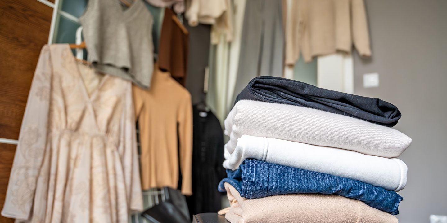 Stacked pile of clothing prepared for resale, donation, or recycling, with wardrobe with outfits in the background. Sustainable fashion choices help reduce waste and support a circular economy.